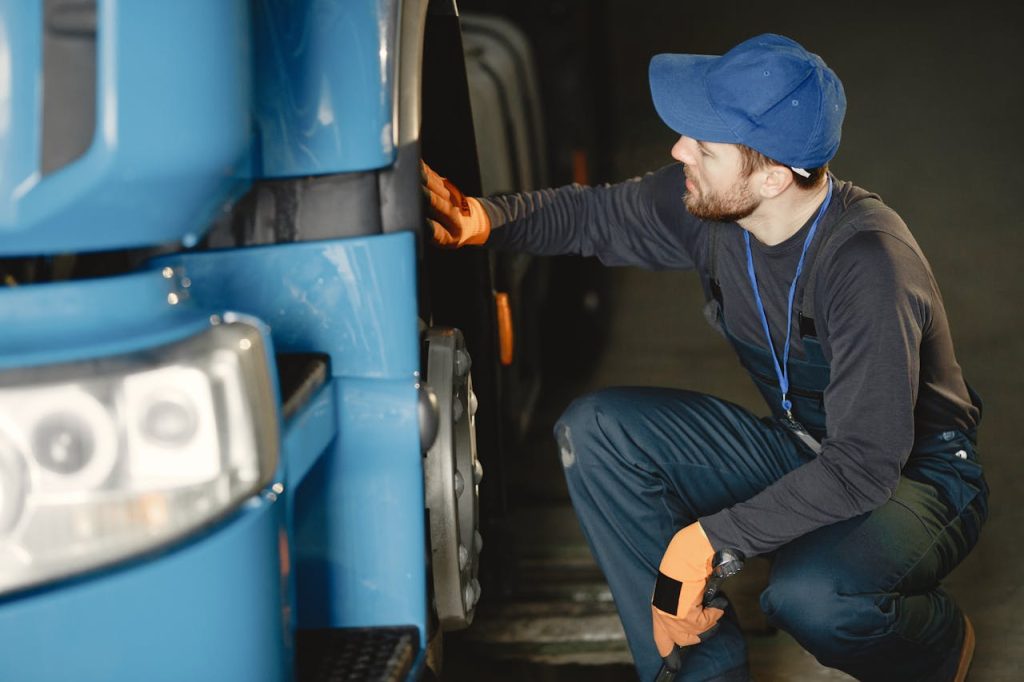 man-in-black-long-sleeve-shirt-and-overall-checking-blue-truck-s-wheel-6720539 Mechanic in uniform checking a blue trucks wheel inside a garage workshop.